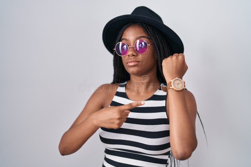 Joven afroamericano con trenzas usando sombrero y gafas de sol con prisa señalando su impaciencia al mirar la cámara fotos de archivo