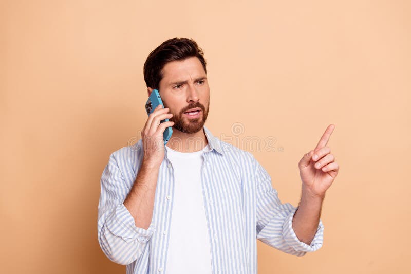 Joven al teléfono con gestos confiados en camisa azul a rayas casuales con fondo beige fotografía de archivo libre de regalías