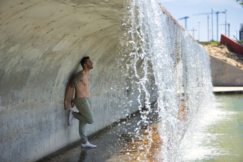 Joven atractivo e hispano sin camisa y con un cuerpo muscular inclinado contra una pared bajo un puente donde una cascada imágenes de archivo libres de regalías