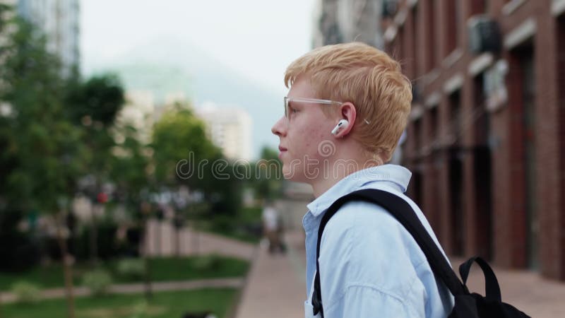 Joven caucásico con gafas y ropa informal caminando en una calle de la ciudad con un edificio de varios pisos como telón de fondo metrajes