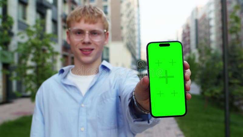 Joven caucásico con gafas y ropa informal mostrando un smartphone con una pantalla verde de burla en una calle de la ciudad almacen de video