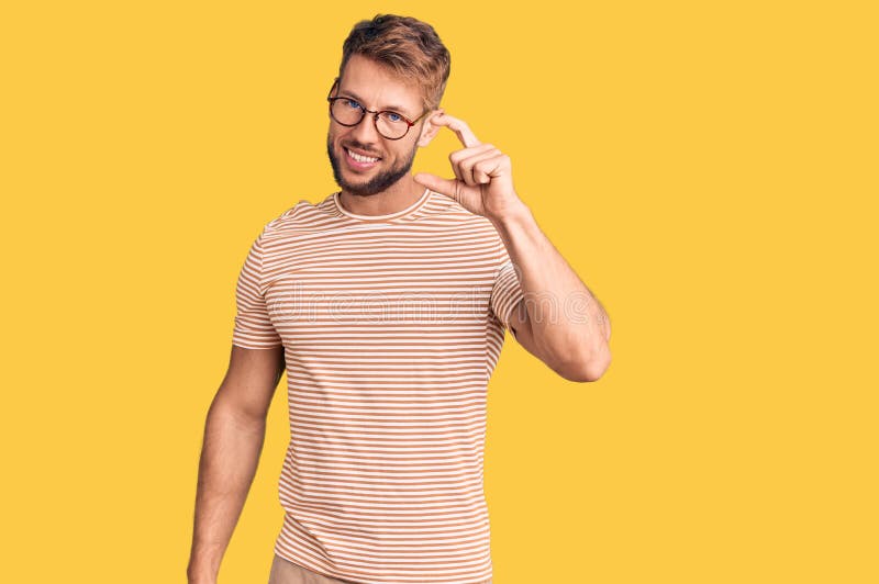 Joven caucásico con ropa informal y gafas sonriendo y gestos confiados con la mano haciendo pequeños signos de tamaño con fotografía de archivo