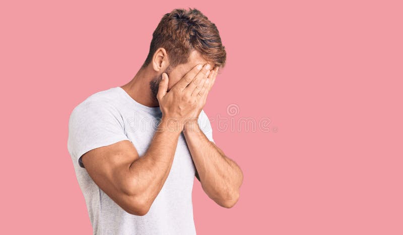 Joven caucásico con una camiseta blanca casual con expresión triste cubriendo la cara con las manos mientras llora imagen de archivo