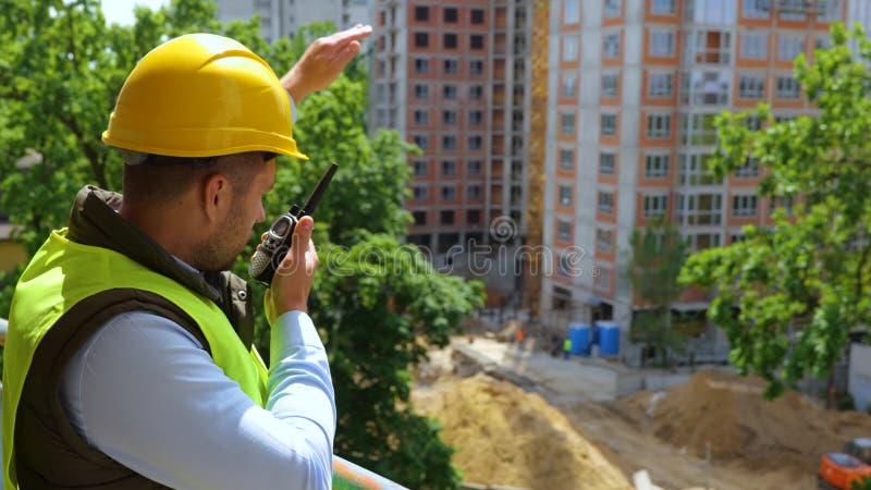 Joven caucásico inspector de construcción con sombrero amarillo duro habla en el proceso de coordinación de construcción de walkie