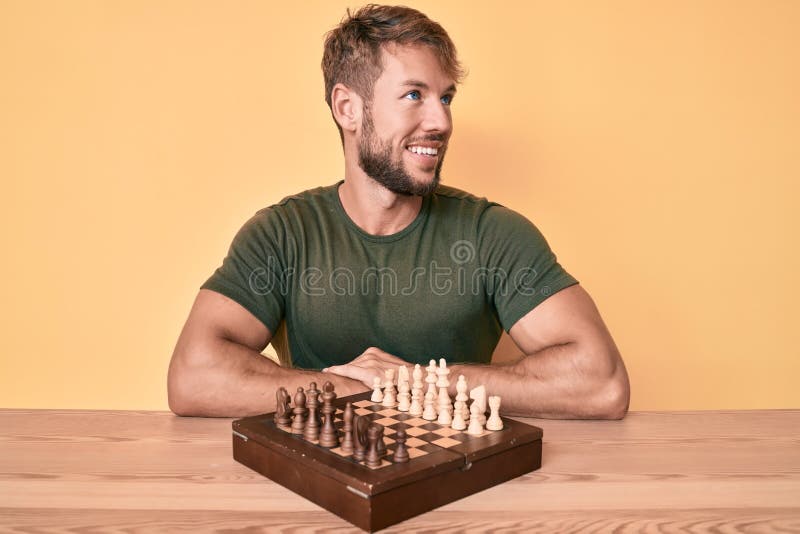 Joven caucásico jugando al ajedrez sentado en la mesa, buscando un lado relajar perfil pose con cara natural y sonrisa segura imagenes de archivo