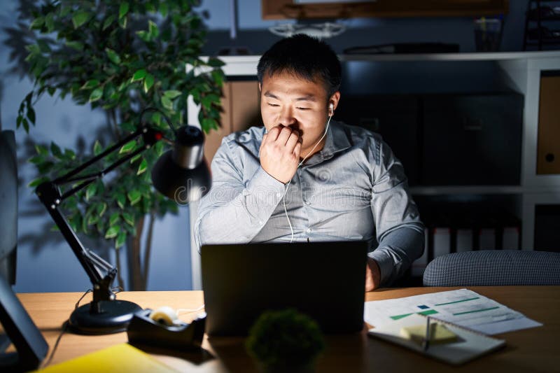 Joven chino que trabaja usando computadora portátil por la noche con aspecto estresado y nervioso con las manos en la boca mordiendo uñas. problema de ansiedad