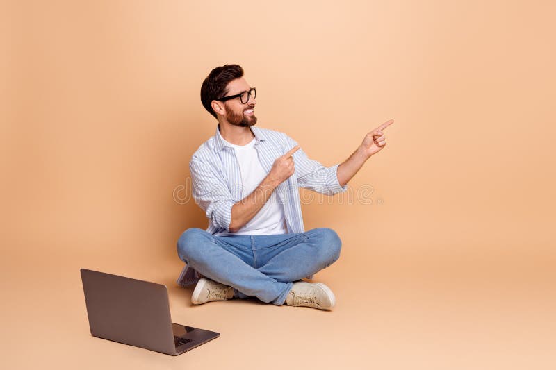 Joven con atuendo casual sentado sobre fondo beige señalando con ambas manos en un gesto promocional imagen de archivo