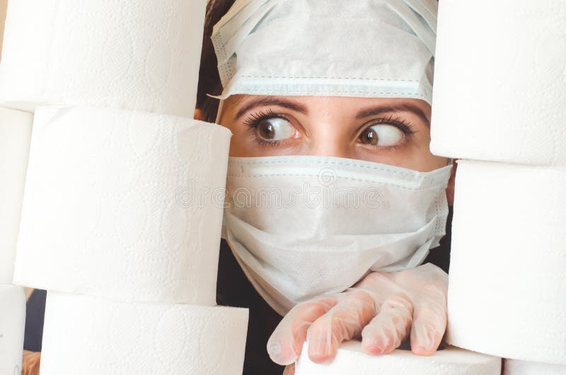 Joven con ojos asustados en dos máscaras faciales de protección contra virus médicos mira a través de pilas de papel higiénico. cu imagen de archivo libre de regalías
