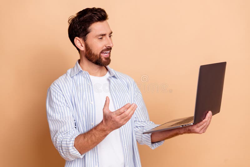 Joven con una camisa casual a rayas sosteniendo una laptop mientras gesta en un fondo beige representando a un hombre confiado y foto de archivo libre de regalías