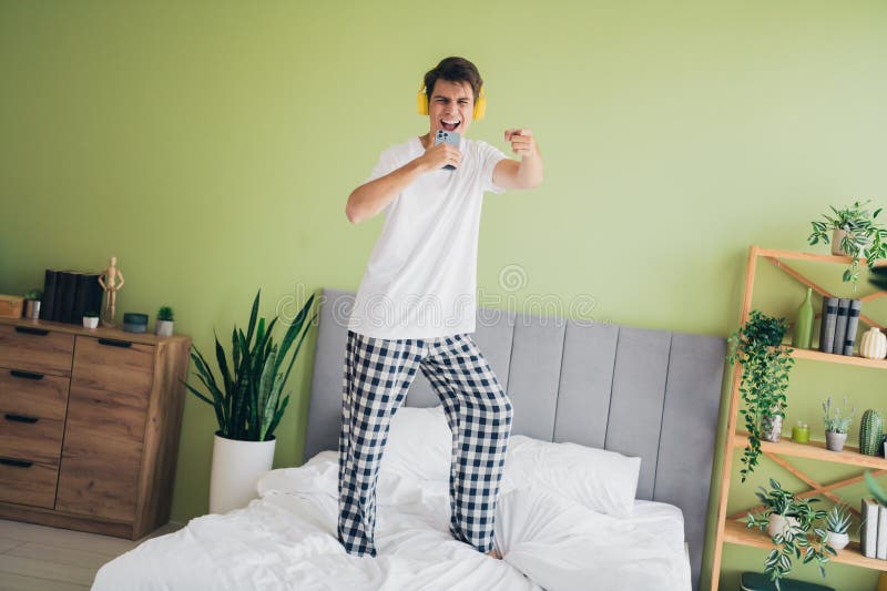 Joven disfrutando de la mañana en pijamas cantando alegremente mientras se pone en cama. Un joven con pijamas y audífonos canta con entusiasmo mientras está de pie sobre una cama en un ambiente acogedor y vibrante de un dormitorio.