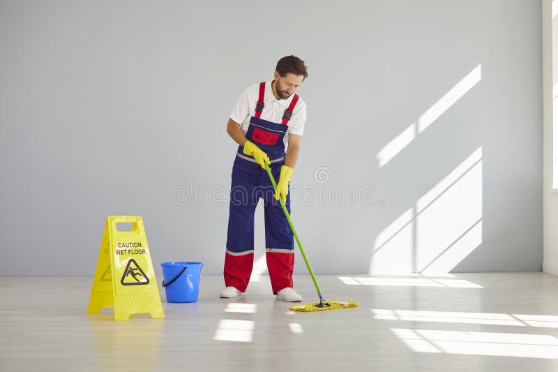 Joven empleado de limpieza en piso de uniforme con señalización de suelo húmedo con precaución en habitación vacía o en la oficina foto de archivo libre de regalías