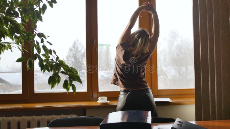 Joven empresaria estira los brazos y el cuello y admira la vista desde la ventana. joven mujer está disfrutando de un soleado