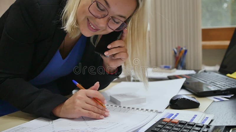 Joven empresaria hablando por teléfono en la oficina examinando los planos en la mesa.