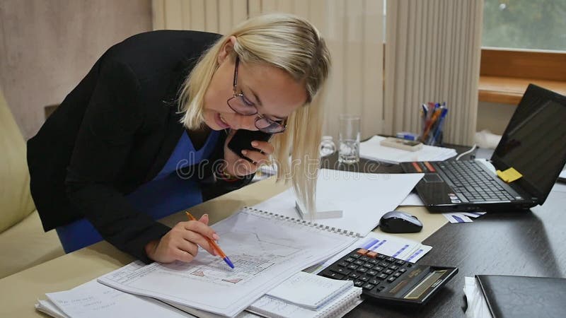 Joven empresaria hablando por teléfono en la oficina examinando los planos en la mesa.