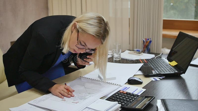 Joven empresaria hablando por teléfono en la oficina examinando los planos en la mesa.