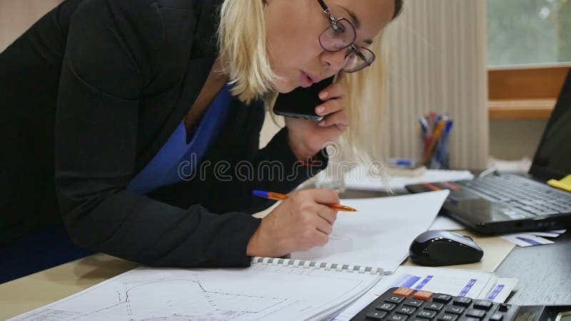 Joven empresaria hablando por teléfono en la oficina examinando los planos en la mesa.