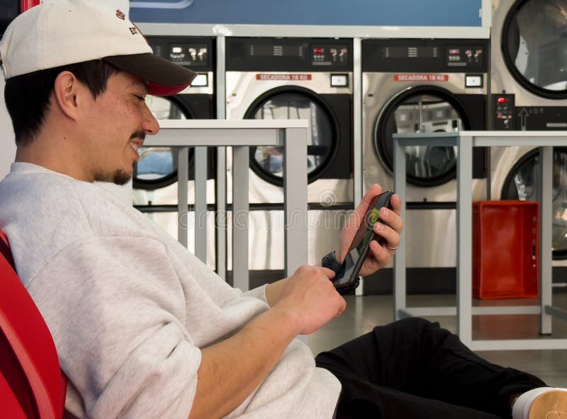Joven entretenido en su celular esperando en la lavandería para lavar su ropa fotografía de archivo