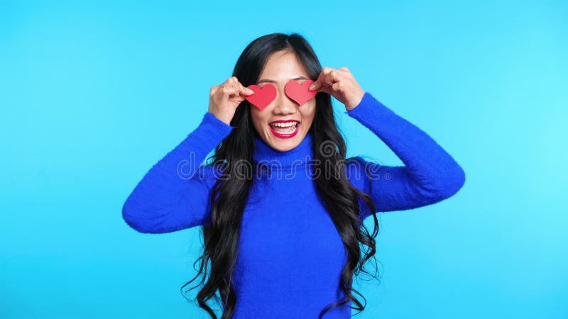 Joven feliz sosteniendo dos corazonitos de papel por sus ojos aislados en fondo azul