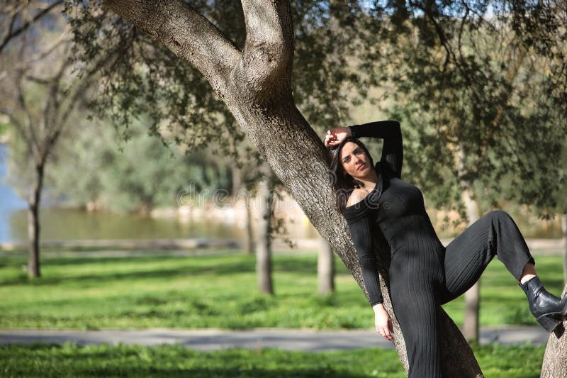 Joven hermosa mujer con traje de salto negro relajado y en calma posando inclinado contra el tronco de un árbol. moda de belleza imagenes de archivo