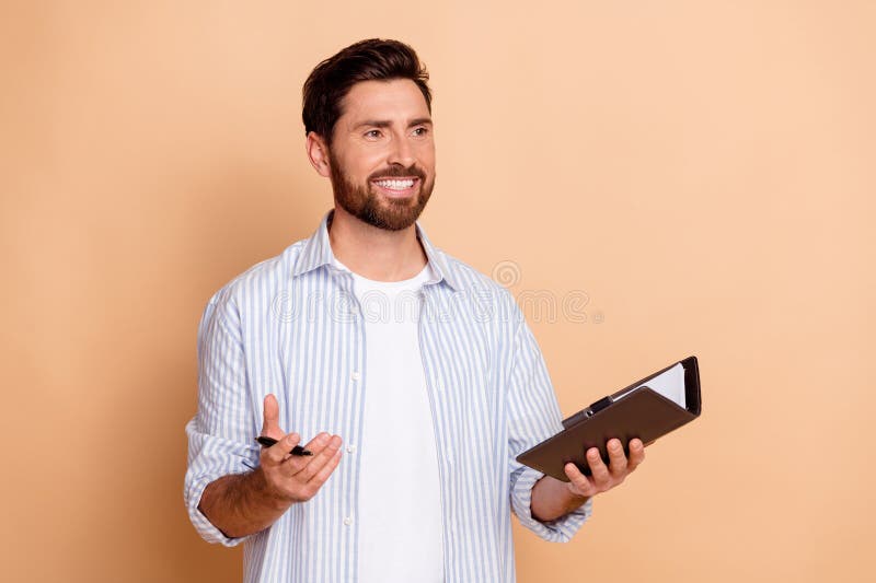 Joven hombre apuesto con una camisa a rayas sujetando el portapapeles y la pluma mientras hacía gestos sobre un fondo beige fotos de archivo libres de regalías