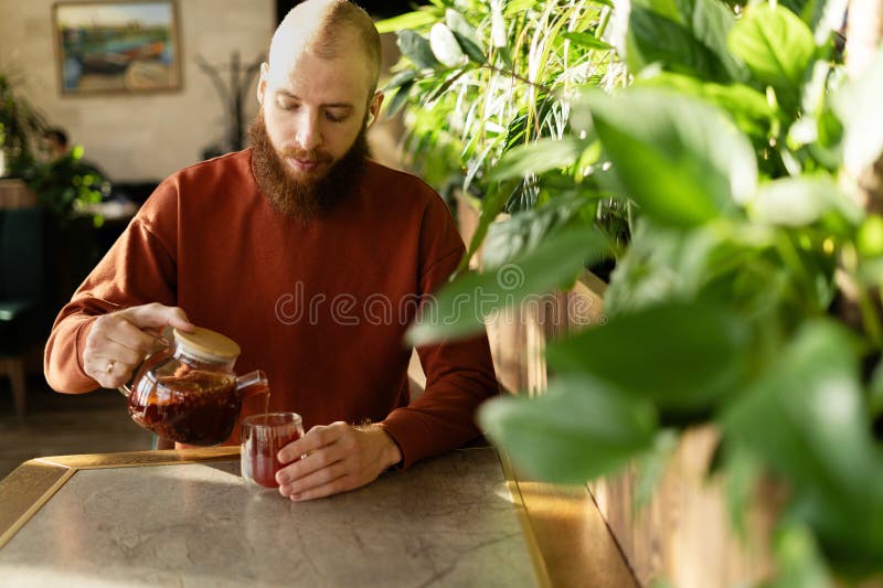 Joven hombre barbudo en un café vertiendo té de fruta en una taza de vidrio fotos de archivo