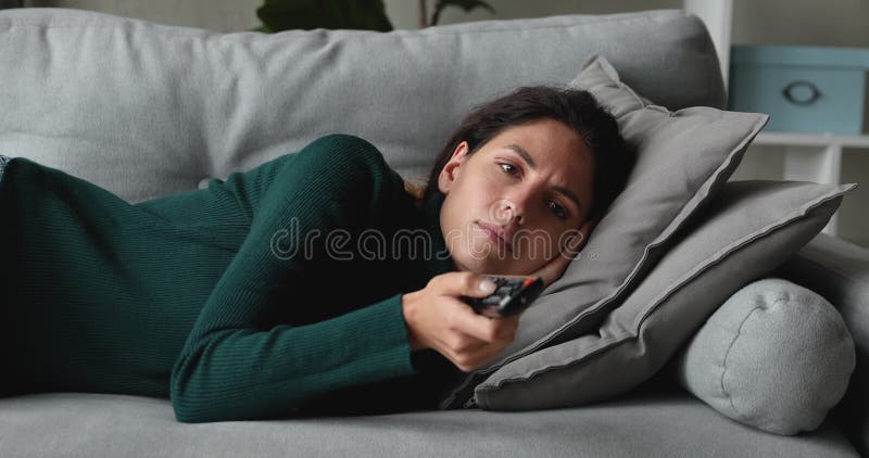 Joven infeliz acostada en el sofá sintiéndose aburrida viendo la tele.