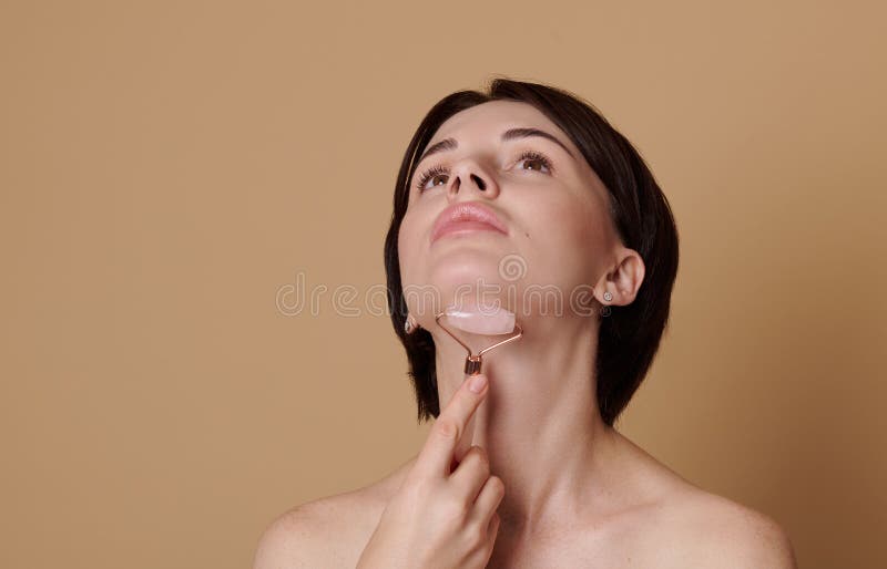 Joven morena haciendo un masaje de elevación en el cuello con un masaje jade roller. retrato de belleza sobre fondo beige con fotos de archivo libres de regalías