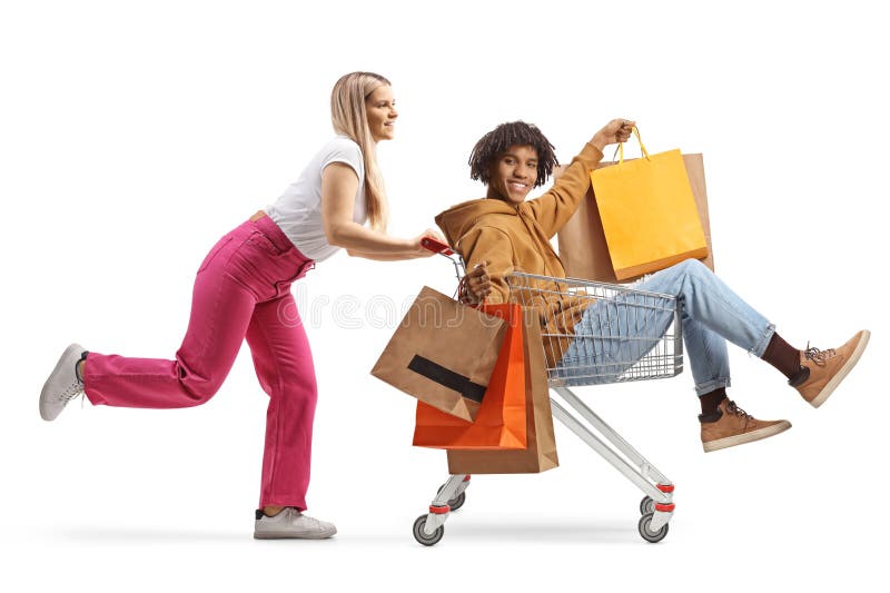 Joven mujer corriendo y empujando a un joven afroamericano dentro de un carrito con bolsas de compras. Mujeres jóvenes corriendo y empujando a un joven afroamericano dentro de un carrito con bolsas de compras aisladas en fondo blanco