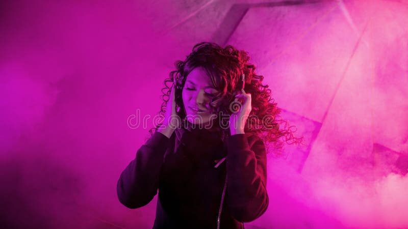 Joven mujer rizada en grandes audífonos sonríe, escucha música, canta, baila, salta, cierra los ojos imágenes de archivo libres de regalías