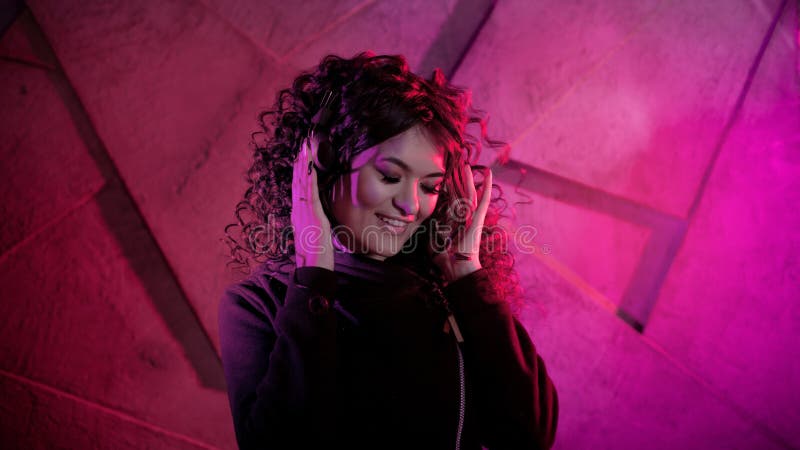Joven mujer rizada en grandes audífonos sonríe, escucha música, canta, baila, salta, cierra los ojos fotografía de archivo