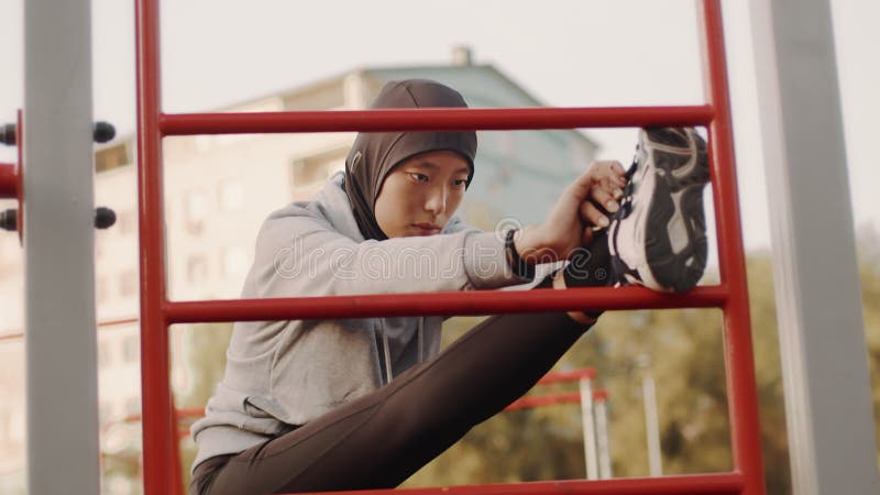Joven musulmana asiática con hijab haciendo ejercicio de estiramiento al aire libre en área de entrenamiento con su pierna en las metrajes