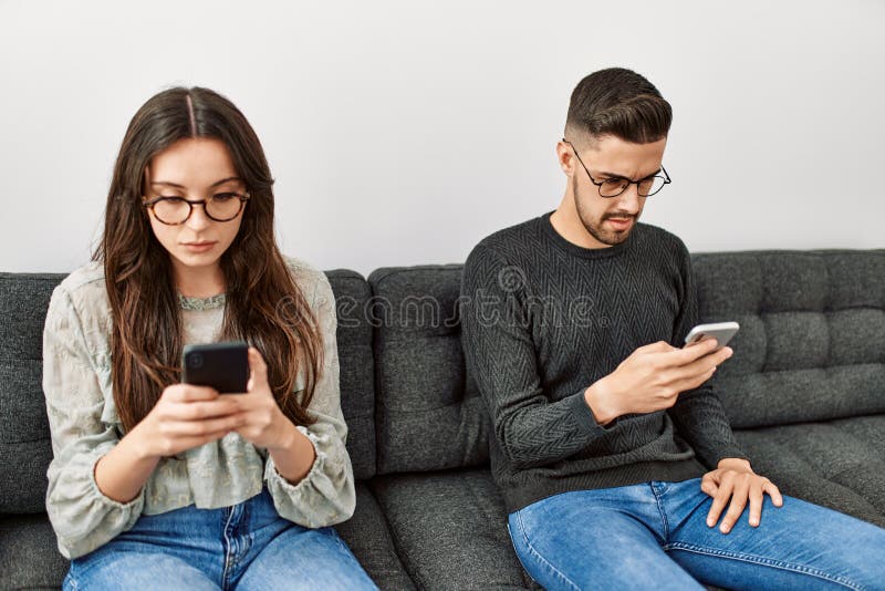 Joven pareja hispana con expresión seria usando un smartphone en casa foto de archivo