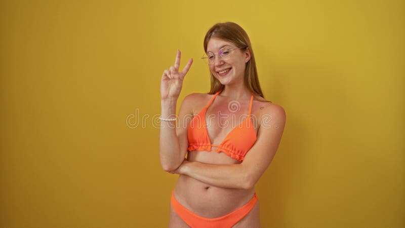 Joven rubia parada en bikini con un cartel de victoria sonriendo alegremente sobre un aislado fondo amarillo mostrando almacen de metraje de vídeo