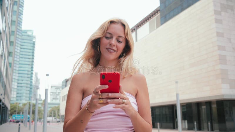Joven rubia sonriente usando el teléfono al aire libre disfrutando de un día soleado usando un traje informal centrado en el móvil metrajes