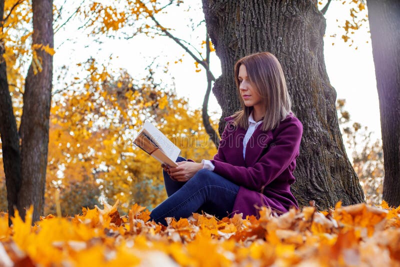 Young woman sits under a tree and reads a paper book. Girl in white shirt and purple coat are sitting on fallen yellow leaves in autumn park. Young woman sits under a tree and reads a paper book. Girl in white shirt and purple coat are sitting on fallen yellow leaves in autumn park.