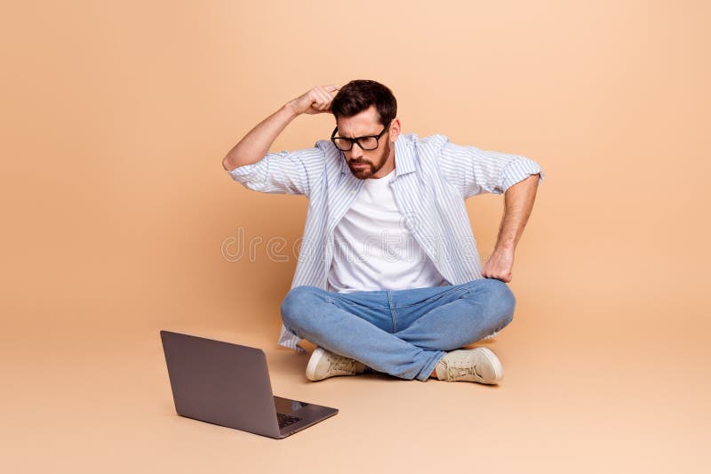 Joven sentado en un fondo beige mientras piensa y mira la laptop imagenes de archivo
