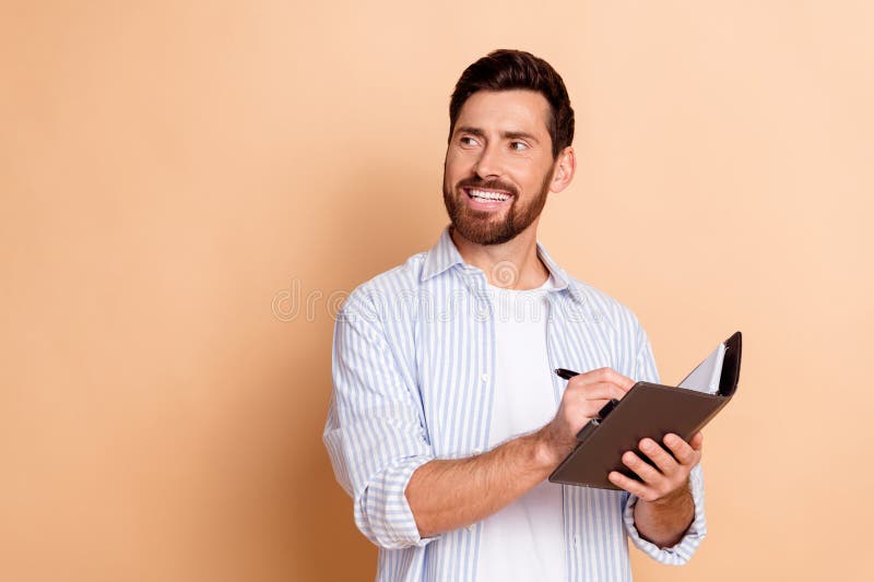 Joven sonriendo mientras escribe en un cuaderno sobre un fondo beige usando una camisa a rayas azules con una moda imagenes de archivo