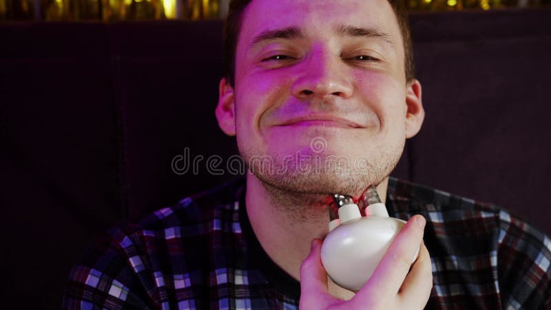 Joven sonriendo mirando la cámara y usando un masajeador 3d para la cara. relajación de los hombres sentados en el sofá y usando e