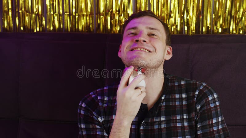 Joven sonriendo y usando un masajeador 3d para la cara. relajado hombre sentado en el sofá y usando un masajeador eléctrico en la