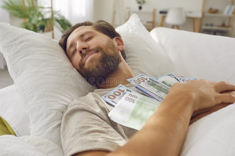 Joven sonriente durmiendo en la cama con un montón de dinero en efectivo manteniendo sus ahorros en casa. imagenes de archivo