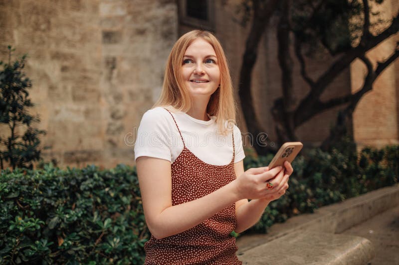 Girl pointing finger on screen smartphone. Young smiling elegant woman holding smartphone walking on street outdoors. Old beautiful city center of Valencia Spain. Girl pointing finger on screen smartphone. Young smiling elegant woman holding smartphone walking on street outdoors. Old beautiful city center of Valencia Spain