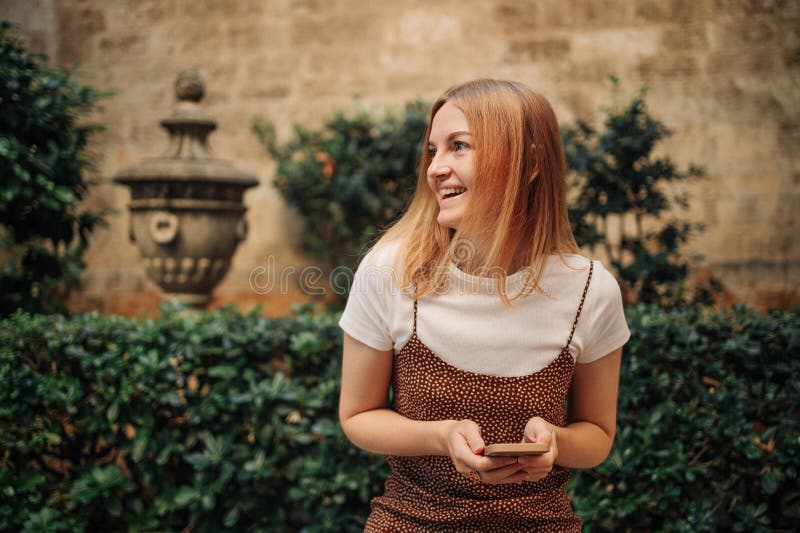 Girl pointing finger on screen smartphone. Young smiling elegant woman holding smartphone walking on street outdoors. Old beautiful city center of Valencia Spain. Girl pointing finger on screen smartphone. Young smiling elegant woman holding smartphone walking on street outdoors. Old beautiful city center of Valencia Spain