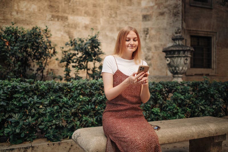 Girl pointing finger on screen smartphone. Young smiling elegant woman holding smartphone walking on street outdoors. Old beautiful city center of Valencia Spain. Girl pointing finger on screen smartphone. Young smiling elegant woman holding smartphone walking on street outdoors. Old beautiful city center of Valencia Spain