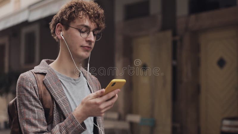 Joven sonriente en lentes con auriculares con chaqueta chaqueta con su Smartphone, mirando a la pantalla Mirando