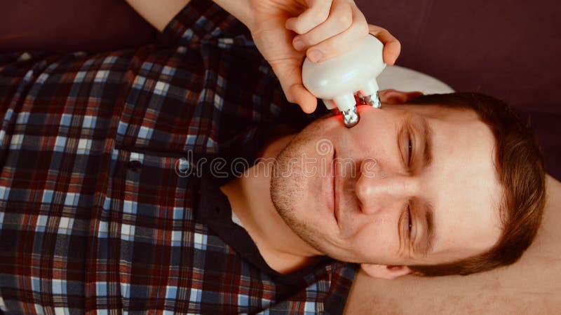 Joven sonriente mirando la cámara y usando un masajeador 3d para la cara. macho relajado tendido en el sofá y usando electricidad