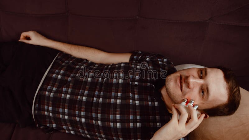 Joven sonriente usando un masajeador 3d para la cara. macho relajado tendido en el sofá y usando un masaje eléctrico. concepto de