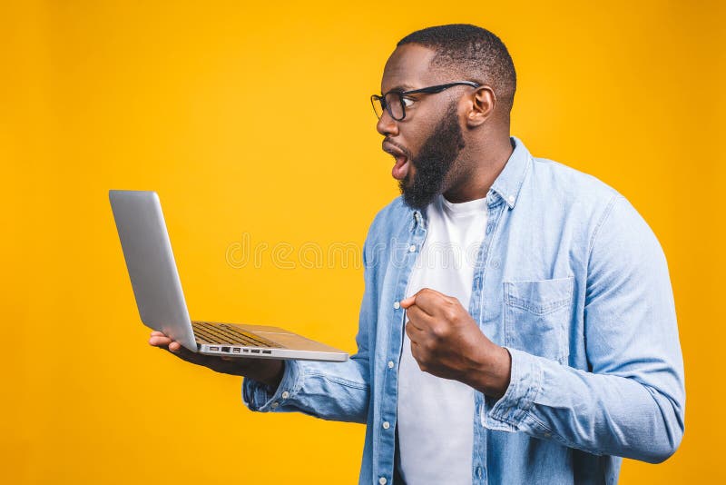 Joven sorprendido hombre africano de pie y usando computadora portátil aislado sobre fondo amarillo fotos de archivo