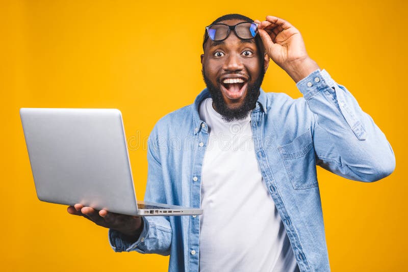 Joven sorprendido hombre africano de pie y usando computadora portátil aislado sobre fondo amarillo fotos de archivo libres de regalías