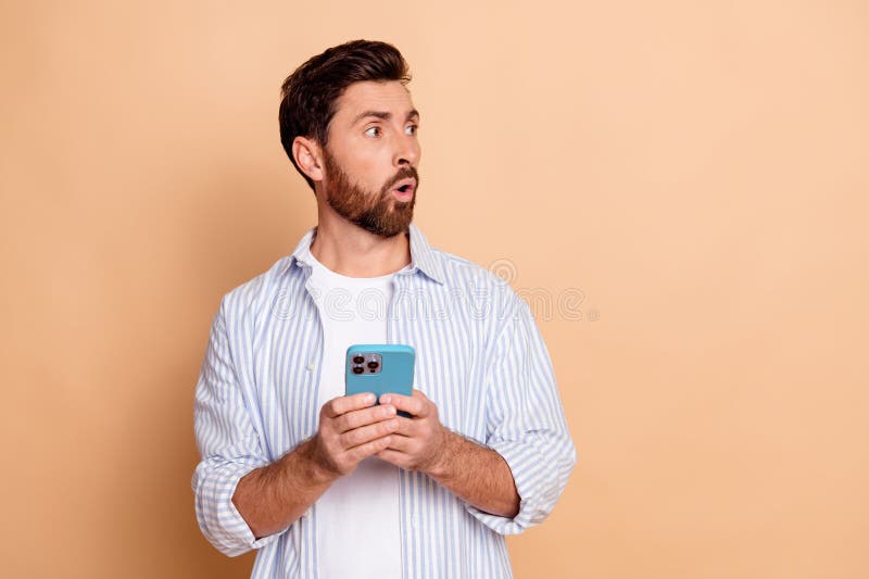 Joven sorprendido sosteniendo un teléfono con una camisa casual de rayas azules contra un fondo beige expresando asombro imagen de archivo libre de regalías