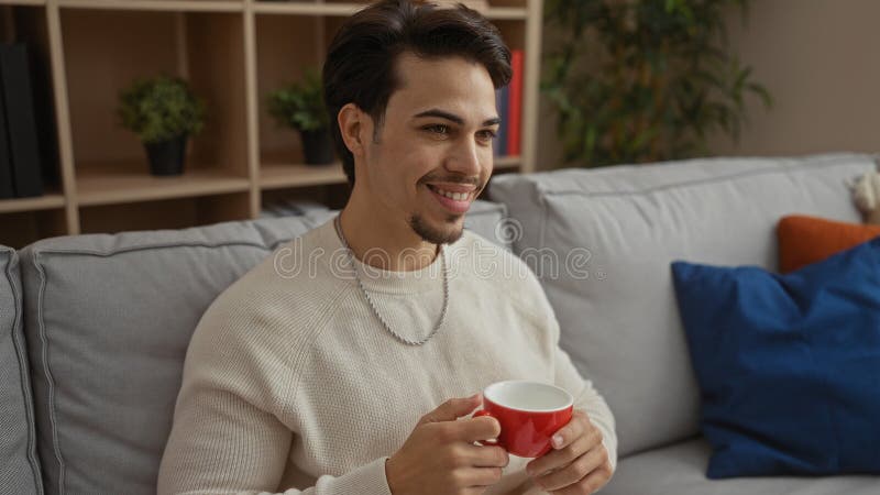 Joven sosteniendo una taza de café sentado en el sofá en un acogedor salón con almohadas y plantas decorativas que crean un hogar fotografía de archivo libre de regalías